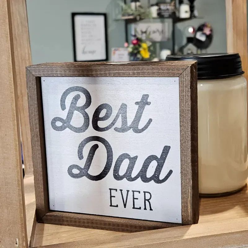 A wooden-framed sign that reads "Best Dad Ever" in black text sits on a shelf next to a large candle in a glass jar. Blurred home decor items and signs are visible in the background.