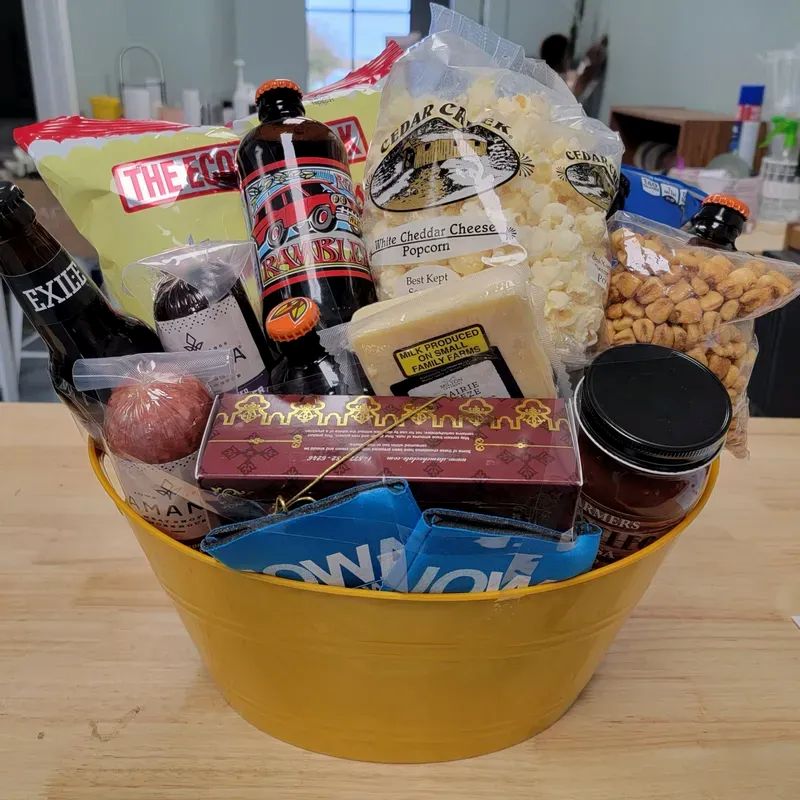 A yellow bucket filled with assorted snacks and drinks, including popcorn, cheese, crackers, beer bottles, sausage, nuts, chocolate, and dips, displayed on a wooden table in a kitchen setting.