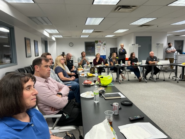 A group of people sits around tables in a brightly lit office meeting room, attentively listening to a presentation. Snacks and drinks are on the tables, and various office items and posters are visible in the background.