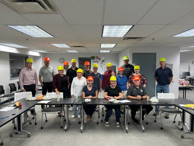 A group of sixteen people wearing colorful hard hats pose for a photo in an office meeting room, some seated and some standing behind a long table with food and drinks.