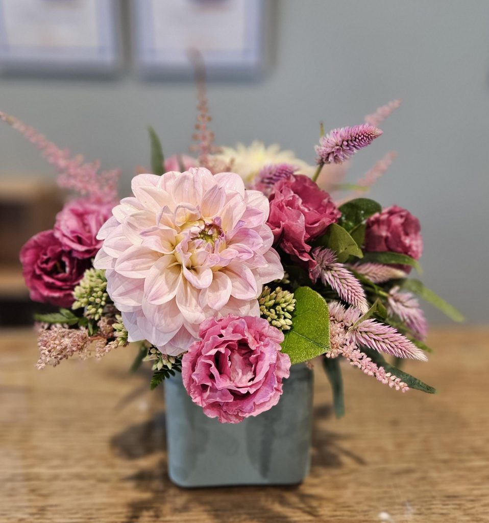 A flower arrangement in a square gray vase featuring pink dahlias, roses, and various purple and green foliage, set on a wooden table with a blurred background.