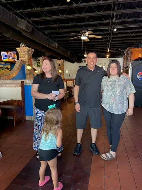 Four people pose inside a restaurant with brick walls and booths. Three adults stand and smile at the camera, while a young girl in a bow and shorts faces them, looking away from the camera.