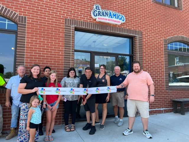 A group of people stands smiling in front of El Gran Amigo Mexican Restaurant, holding a ribbon for a ribbon-cutting ceremony on a sunny day.