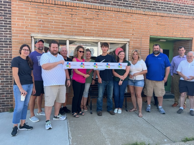 A group of people stands outside a brick building, holding a ribbon for a ribbon-cutting ceremony. Some are smiling and posing, with one person holding scissors in the center.