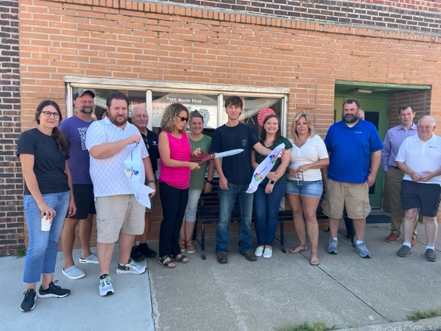 A group of people stands in front of a brick building. A woman in pink holds large scissors, cutting a ribbon, while others smile and watch, celebrating a ribbon-cutting ceremony on a sunny day.