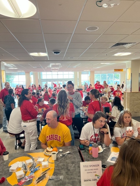 A large group of people, mostly wearing red and white shirts, gather in a cafeteria-style room. Some are seated at tables, eating and talking, while others stand and mingle. The atmosphere is lively and social.