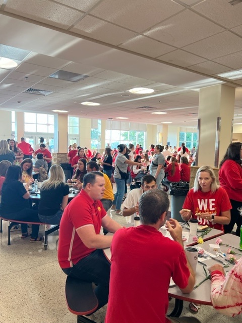 Large group of people, mostly wearing red shirts, sit and stand around tables in a bright, spacious cafeteria, engaged in conversation and eating. Sunlight streams in through large windows in the background.
