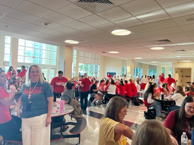 A large group of people, many wearing red shirts, gather in a brightly lit cafeteria with round tables. Some are seated and talking, while others stand and mingle. A woman in white pants stands smiling in the foreground.