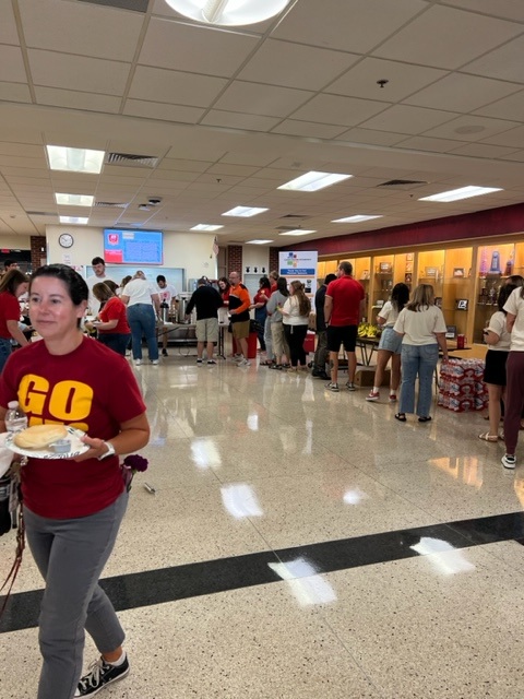 People are lined up in a school cafeteria or lobby area, some getting food. A woman in a red "GO" shirt carries a plate of food and smiles in the foreground. The area is crowded and brightly lit.