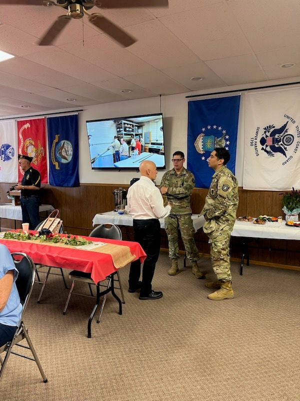 Three people, including two in military uniforms and one in a white shirt, talk in a decorated room with military flags, tables with red tablecloths, and food. A screen displays an image in the background.