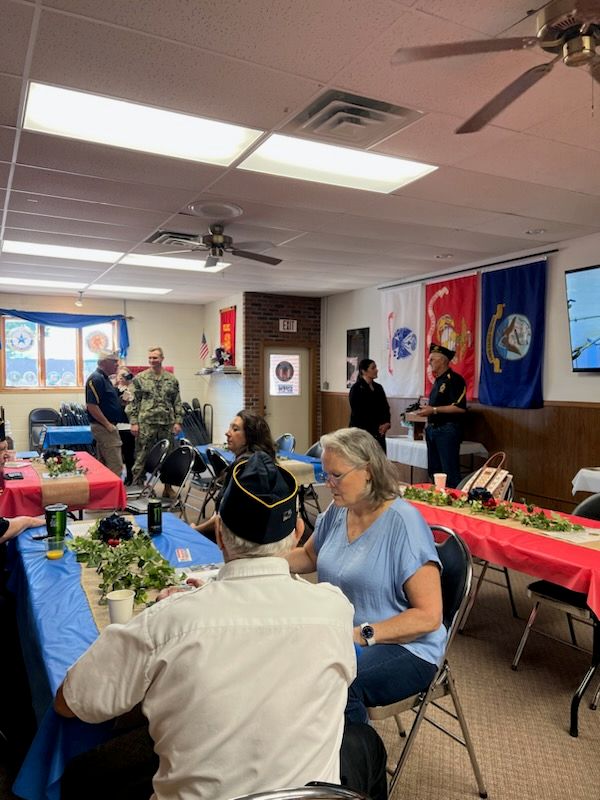 People gathered in a decorated room with military flags on the wall, sitting at tables with red and blue tablecloths. Some are talking, and others are seated, suggesting a social or commemorative event.