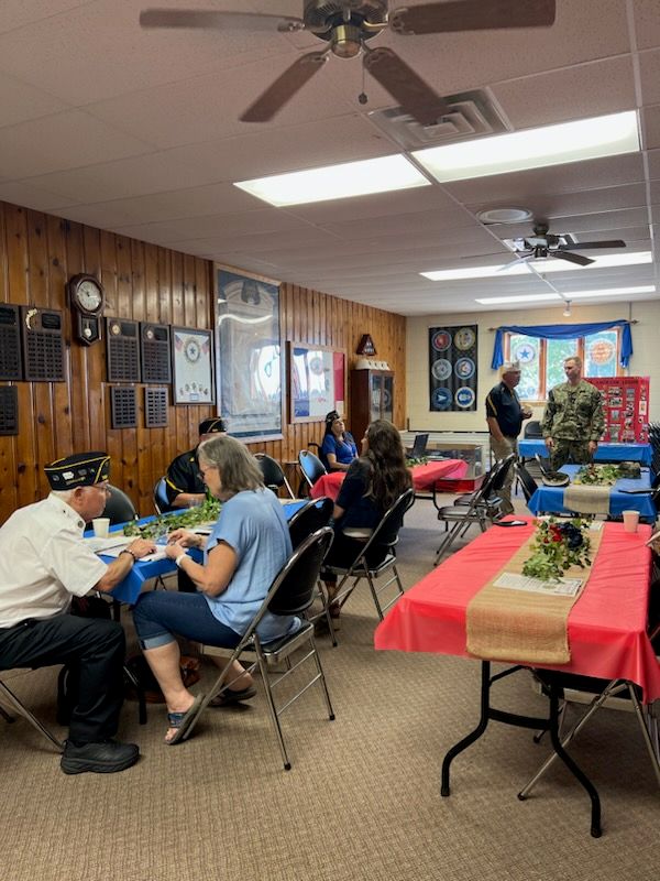 People are sitting and talking at decorated tables in a wood-paneled room with military memorabilia and plaques on the walls. A few people, including someone in a military uniform, are standing and conversing.