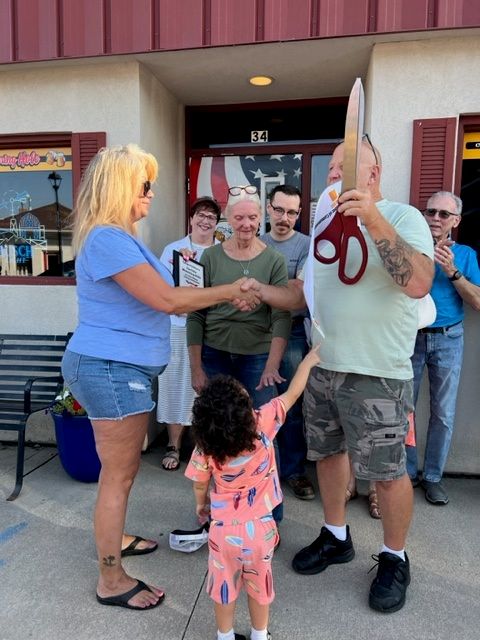 A group of people stands outside a building for a ribbon-cutting ceremony. A woman shakes hands with a man holding large scissors, while a small child reaches toward them. Others smile and clap in the background.