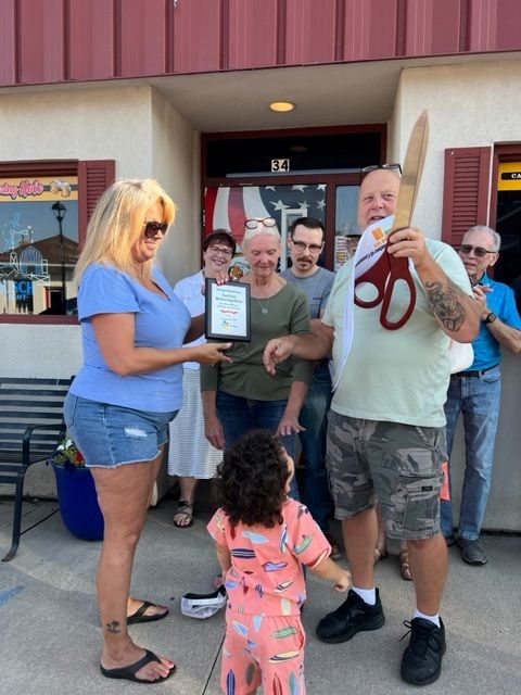 A group of people stand outside a building. A woman hands a plaque to a man holding large novelty scissors. A young child in colorful clothes faces them. Others in the background smile and watch the moment.