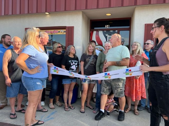 A group of people gather outside a building for a ribbon-cutting ceremony. Several are smiling, and one man holds large scissors, ready to cut the ribbon. The scene looks cheerful and celebratory.