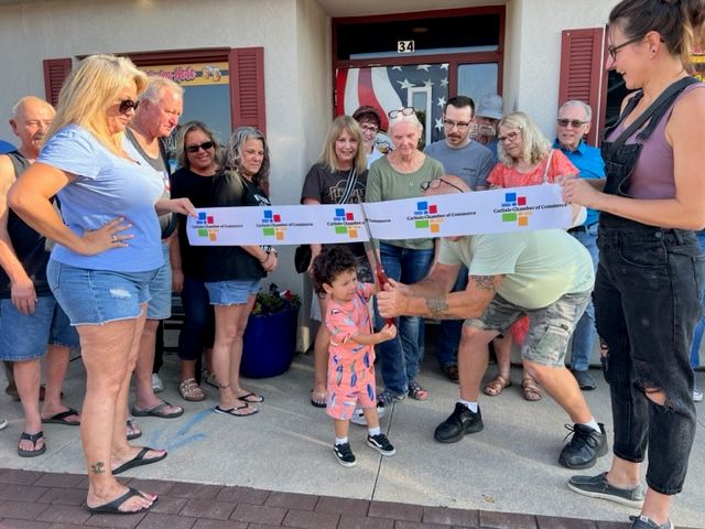 A group of people gathers outside a building for a ribbon-cutting ceremony. An adult helps a young child cut a ribbon, while others smile and watch. The ribbon has colorful logos on it.
