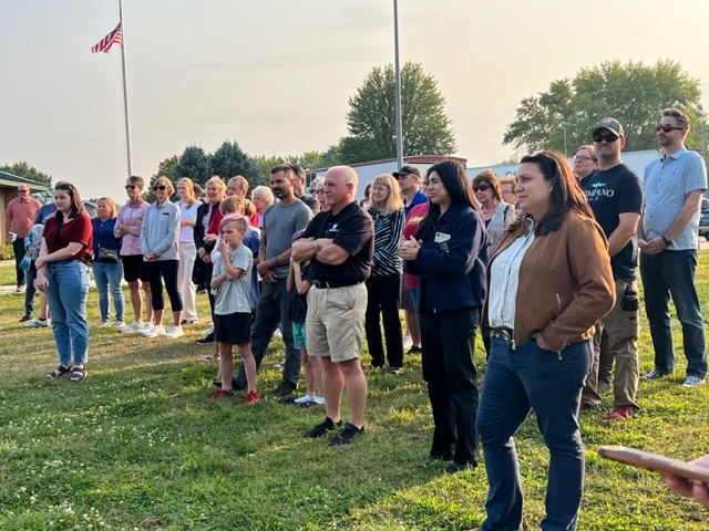 A group of people, including adults and children, stand outside on grass, attentively watching something off-camera. The American flag is visible in the background, along with trees and a clear sky.