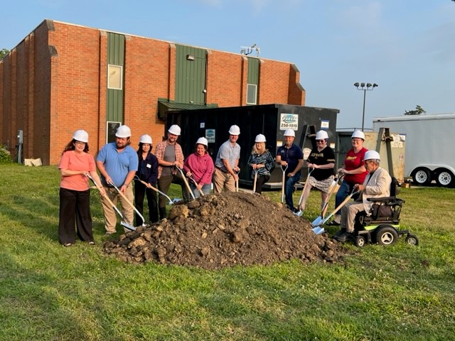 A group of people wearing hard hats stand in a line, each holding a shovel and posing in front of a mound of dirt, with a brick building and trailers in the background on a sunny day.