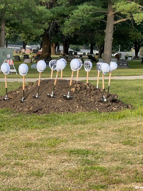 A row of shovels with white hard hats on their handles stands upright in a pile of dirt, set in a grassy area with trees and a cemetery in the background.