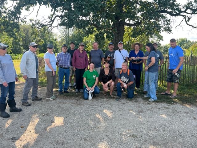 A group of fifteen people, dressed casually, stand and kneel together outside on a gravel path under a large tree with green leaves. A wooden fence and greenery are visible in the background on a sunny day.