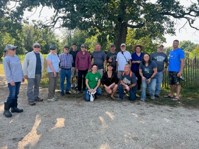 A group of fifteen adults pose together outdoors on a sunny day, standing and kneeling on a gravel path in front of a large tree and a rustic fence, surrounded by greenery.