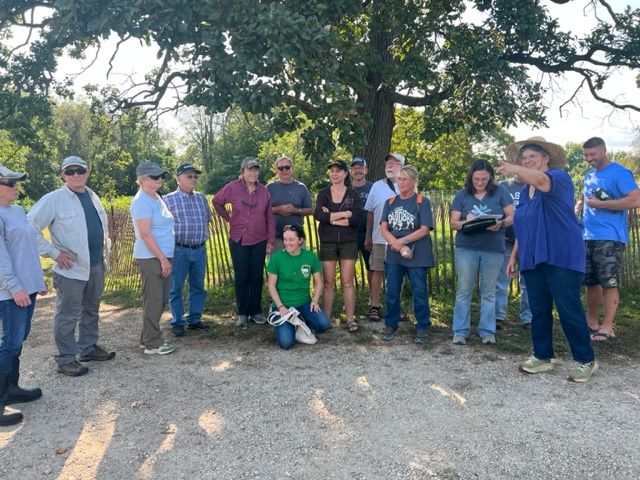 A group of people stands outdoors on a sunny day, some smiling and talking, with trees and a wooden fence in the background. One person in a blue shirt gestures while others listen or watch.