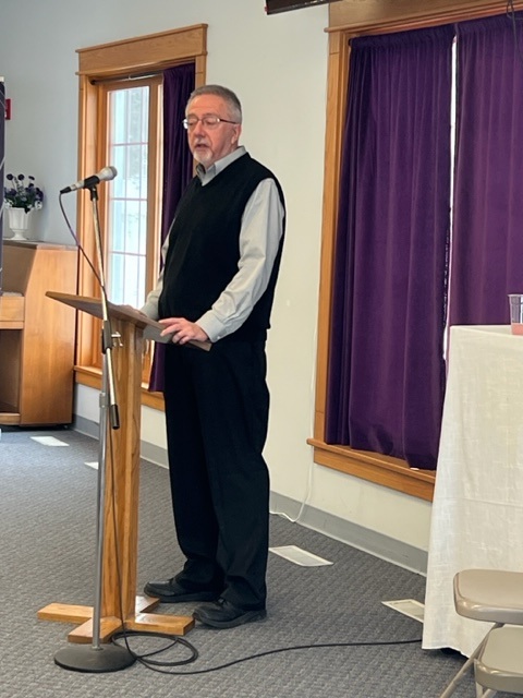 A man in a black vest and dress pants stands at a wooden podium, speaking into a microphone in a room with purple curtains and large windows.
