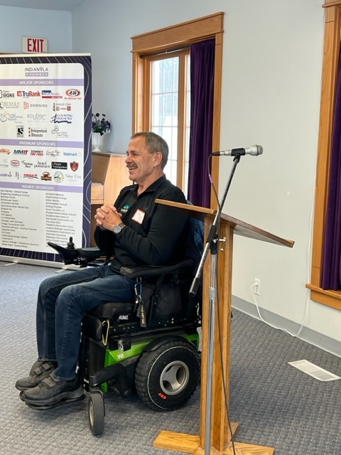 A man in a motorized wheelchair smiles while sitting beside a wooden podium with a microphone in a well-lit room. Behind him is a window and a poster displaying various sponsor logos.