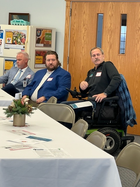 Three men sit at a conference table. One man is in a wheelchair. Papers and a small flower arrangement are on the table. A refrigerator with notices and double wooden doors are visible in the background.