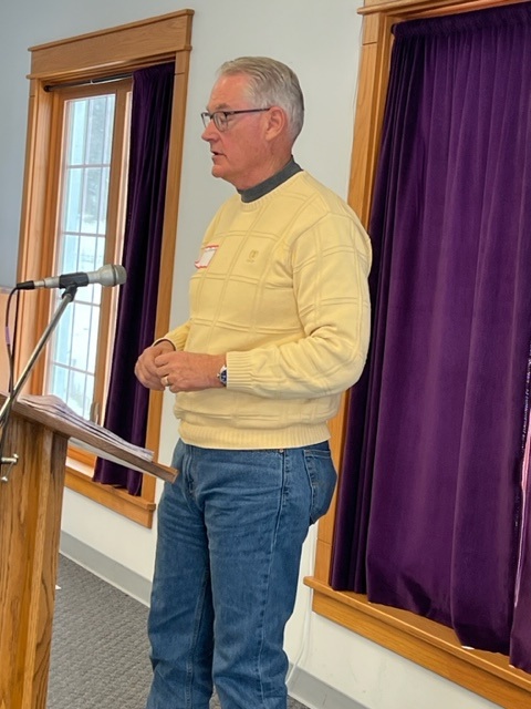 A man with gray hair, glasses, and a yellow sweater stands at a podium speaking into a microphone in a room with purple curtains and large windows.