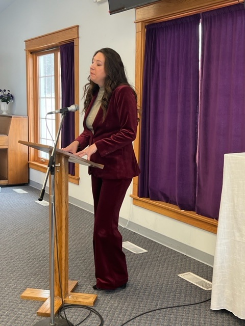 A woman in a burgundy suit stands at a wooden podium, speaking into a microphone in a room with purple curtains and natural light coming through the windows.