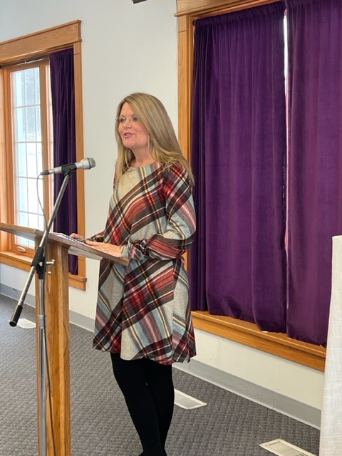 A woman with long blonde hair, wearing a plaid dress and black leggings, stands at a podium and speaks into a microphone in a room with purple curtains and large windows.