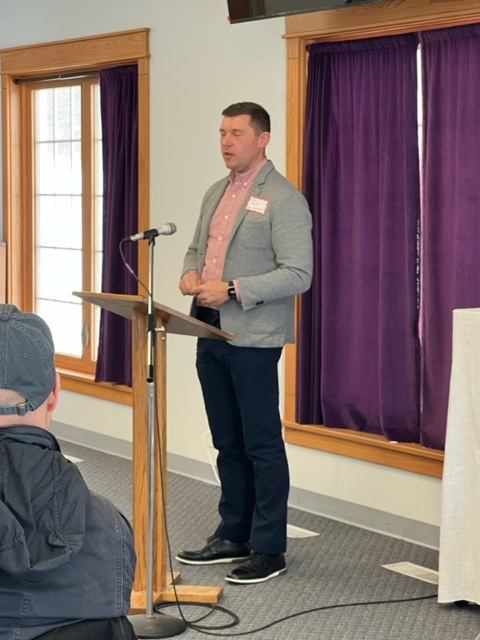 A man in a gray blazer and dark pants speaks at a wooden podium with a microphone in a room with large windows and purple curtains. People are seated and listening to him.