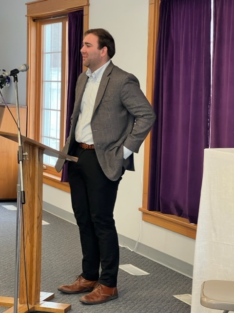 A man wearing a blazer, dress shirt, and slacks stands at a podium, speaking in a room with purple curtains and large windows. A microphone is attached to the podium.