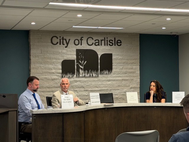 Three people sit at a curved desk in front of a wall reading "City of Carlisle." Two men are on the left, one woman is on the right. Nameplates are on the desk, and the room has a modern design with teal and gray colors.