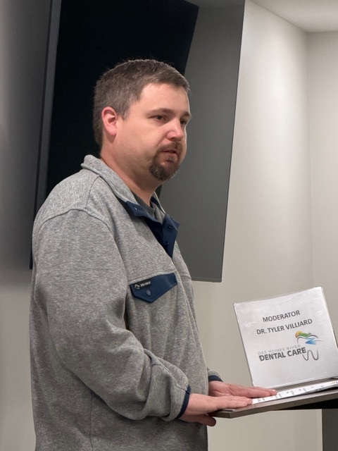 A man in a gray pullover stands at a lectern, looking ahead. The lectern holds a sign that reads "Moderator Dr. Tyler Villiard" and displays a dental care logo. A large screen is mounted on the wall behind him.