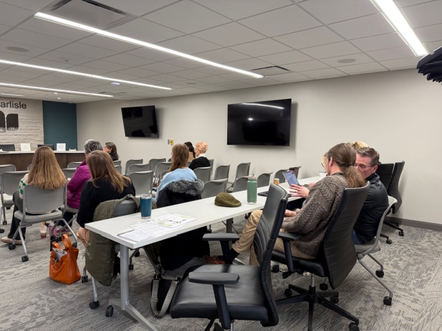 A group of people sit in a modern conference room with gray chairs, facing the front where two large monitors are mounted on the wall. Some attendees are talking, using laptops, or looking forward.