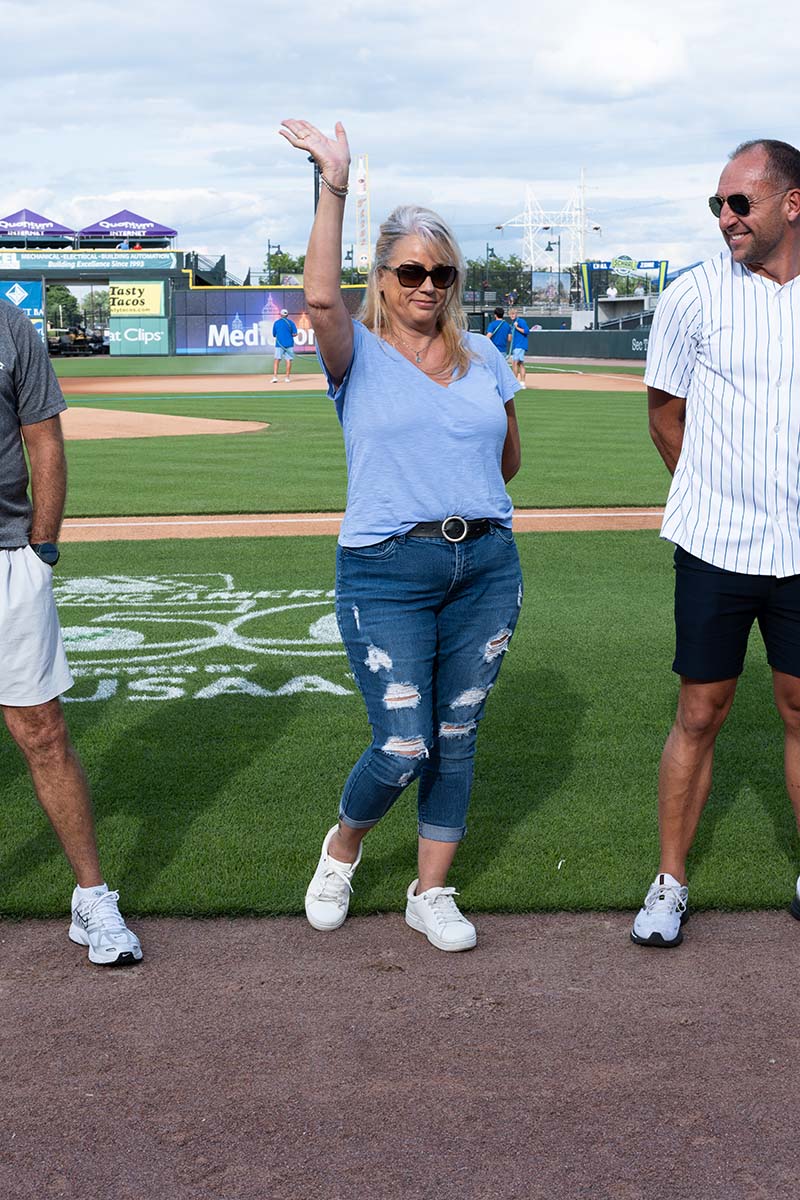 A woman in sunglasses, a light blue shirt, and ripped jeans stands on a baseball field, smiling and waving, flanked by two men, one in white shorts and the other in a striped shirt.