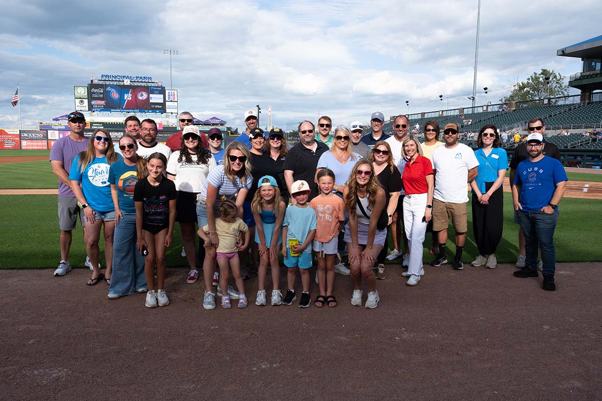 A large group of adults and children pose and smile together on a baseball field, with stadium seats and a scoreboard visible in the background under a partly cloudy sky.