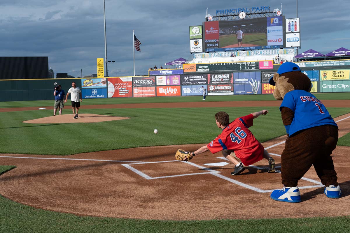 A baseball player in a red jersey reaches to catch a pitch at home plate, while a mascot in a blue jersey stands nearby; people and a scoreboard are visible in the background on a sunny day.