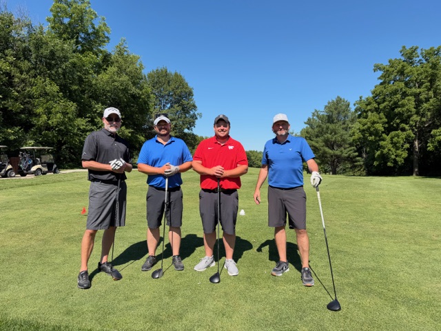 Four men pose on a sunny golf course, each holding a golf club. They are dressed in shorts, polo shirts, and caps, with trees and a golf cart in the background under a clear blue sky.