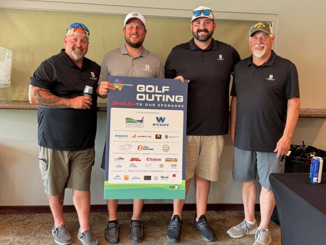 Four men in black golf shirts and caps stand indoors, smiling, with their arms around each other. They are posing next to a large sign thanking sponsors for a golf outing event.