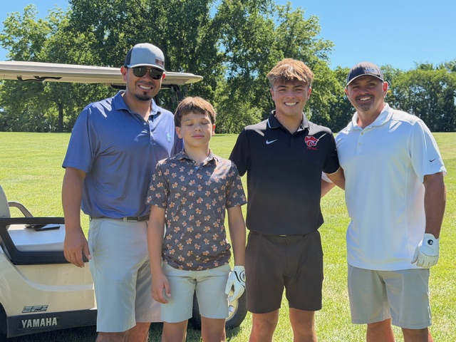 Four people stand together outdoors on a golf course near a golf cart, smiling at the camera. Three adults wear polo shirts and one boy wears a patterned button-up shirt and shorts. Trees and grass are in the background.
