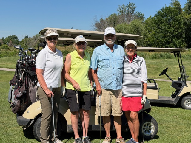 Four adults, three women and one man, stand smiling with golf clubs in front of two golf carts on a sunny day. They are dressed in golf attire on a green course with trees in the background.