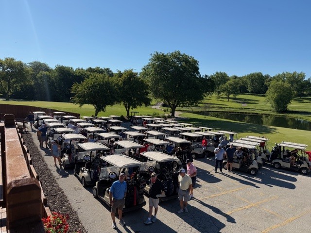 A group of people gather around numerous golf carts in a parking area near a green golf course with trees and a pond visible under a clear blue sky.
