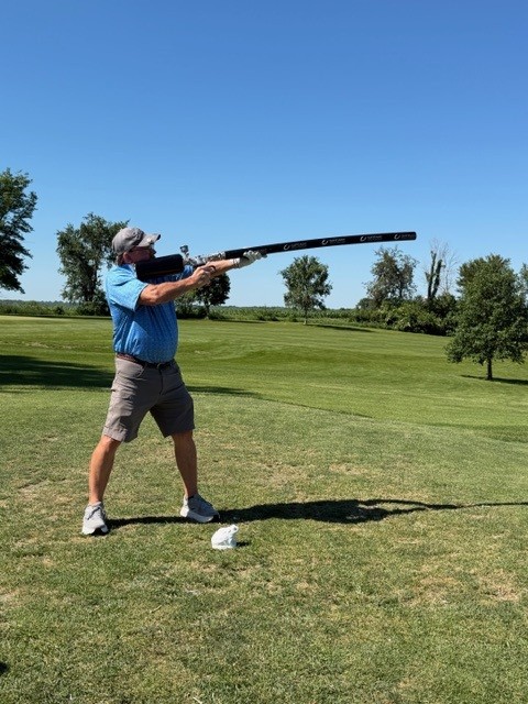 A man in a blue shirt and gray shorts stands on a grassy field, aiming a long, oversized golf club towards the sky on a sunny day with clear blue skies and trees in the background.