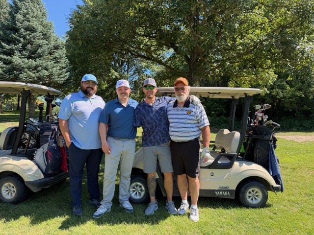 Four men stand together smiling in front of two golf carts on a sunny day, with trees and grass in the background. They are dressed in golf attire and appear to be enjoying a day on the golf course.