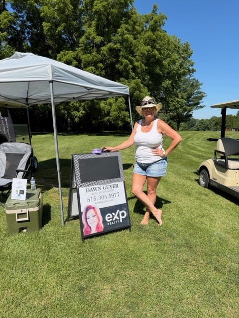 A woman in a white tank top, denim shorts, and a hat stands next to a sign for Dawn Guyer, eXp Realty, on a grassy area with a canopy tent and golf cart nearby under a sunny sky.