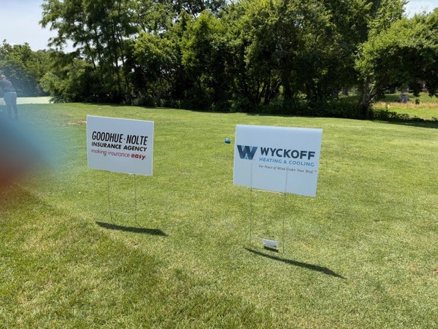 Two business signs on a grassy lawn, one for Goodhue-Nolte Insurance Agency and the other for Wyckoff Heating & Cooling, with trees and sunlight in the background.