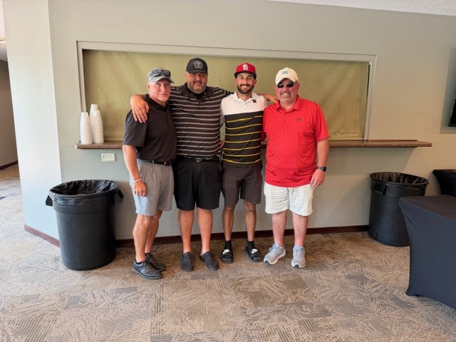 Four men stand together indoors, smiling at the camera. They are dressed in golf attire, wearing hats and shorts. Two trash bins and a counter with stacked cups are visible in the background.